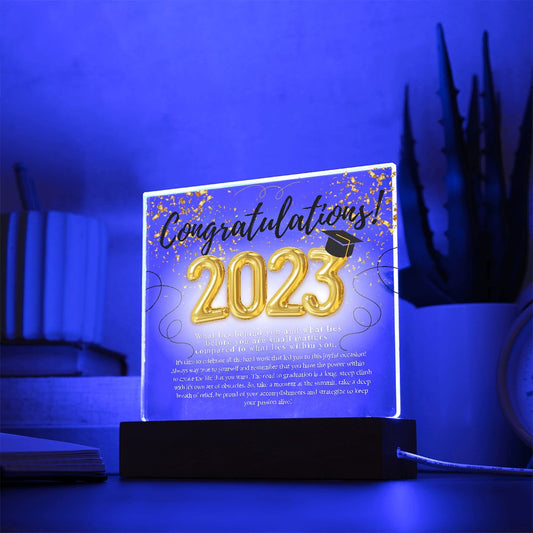 A sentimental plaque with heartfelt message, for anyone who graduated sitting on top of an illuminated base, placed on a desk with plants, books, pens, notebook, and a coffee cup.